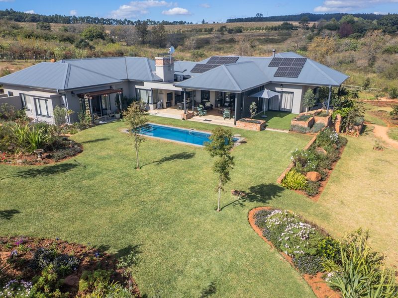 Aerial view of a modern single-story residence featuring a swimming pool, covered patio, solar panels, and extensive landscaped gardens in a rural setting.