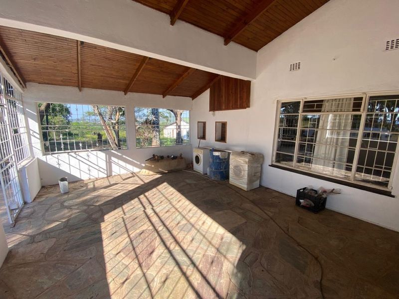 A spacious covered patio featuring exposed timber ceilings, stone flooring, and large windows with security bars, offering a versatile outdoor entertainment area.