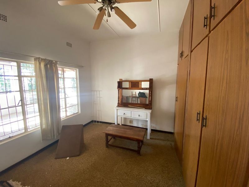 A bedroom featuring built-in wooden cupboards, a ceiling fan, and large windows with security bars, requiring cosmetic updates to the flooring and finishes.