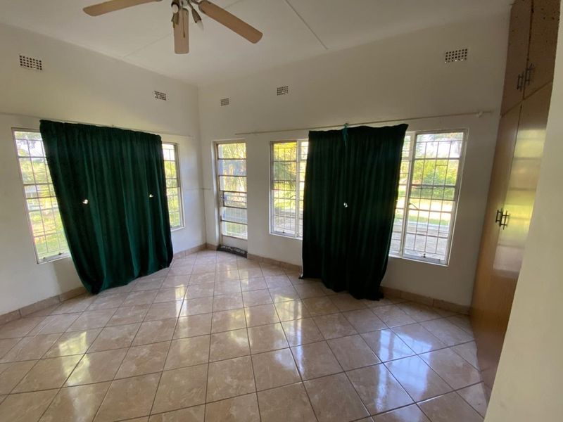 A spacious, empty bedroom featuring tiled flooring, built-in cupboards, a ceiling fan, and large windows with security bars and a security gate.