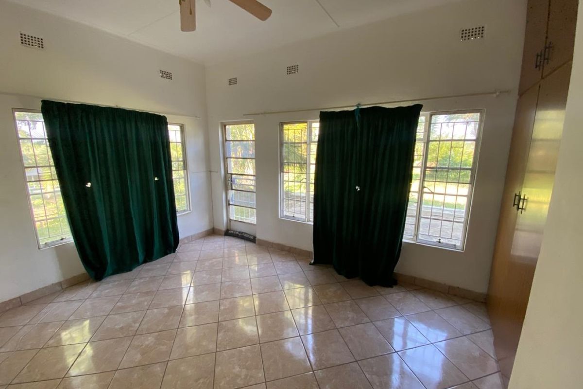 A spacious, empty bedroom featuring tiled flooring, built-in cupboards, a ceiling fan, and large windows with security bars and a security gate.