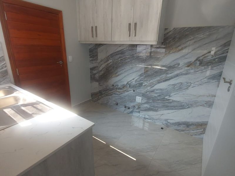 A modern, empty kitchen area featuring marble-look tiled splashbacks, stone-look countertops, a double bowl stainless steel sink, and ample upper cabinetry.