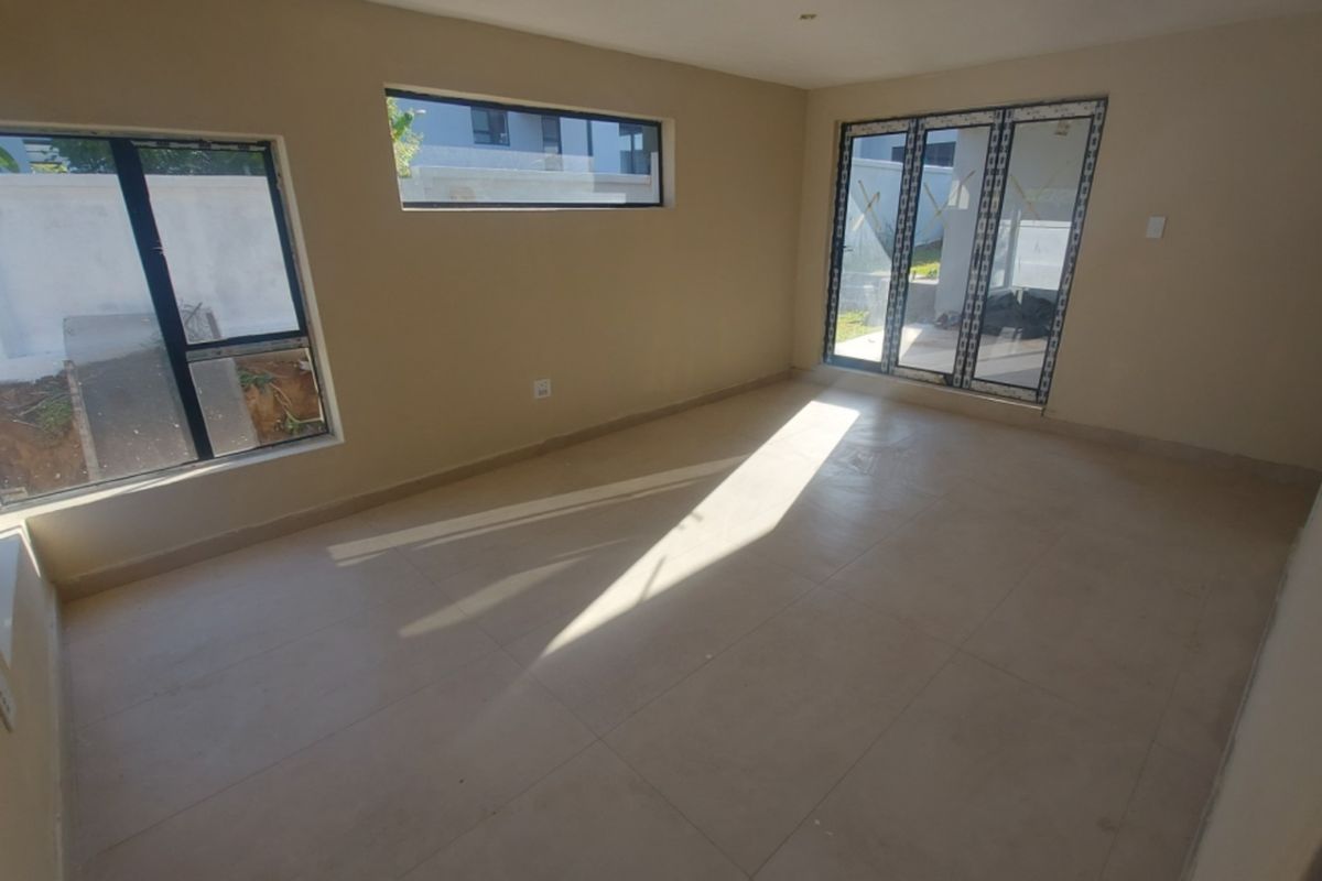 A bright, empty living room featuring neutral tiled flooring, modern aluminium window frames, and sliding doors leading to the exterior.