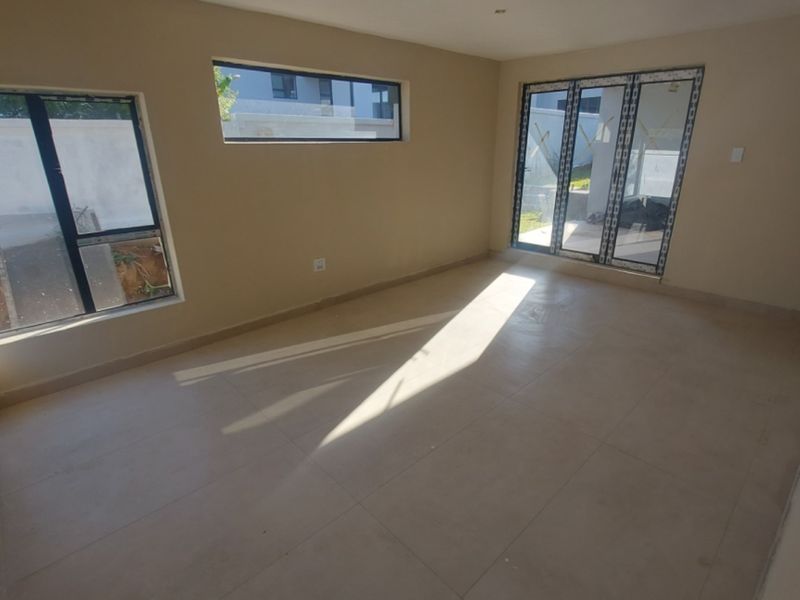 A bright, empty living room featuring neutral tiled flooring, modern aluminium window frames, and sliding doors leading to the exterior.