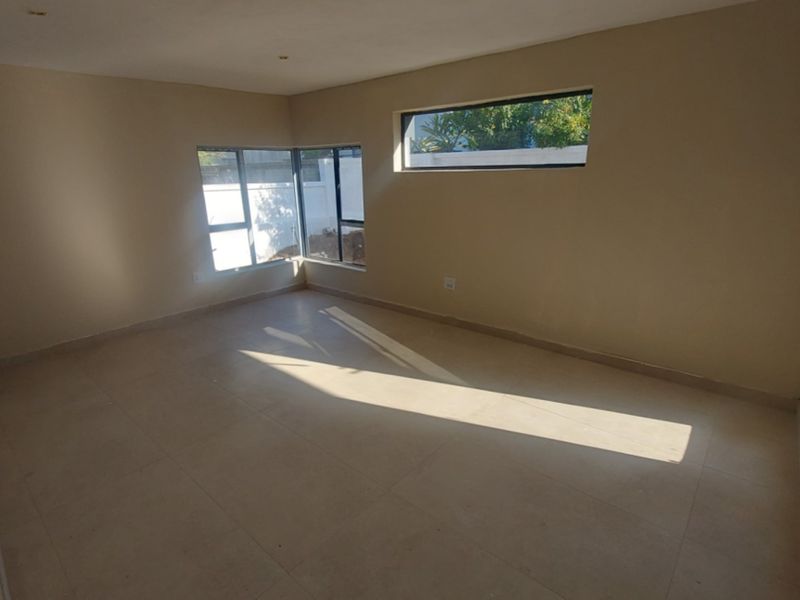 A bright, empty living room featuring neutral tiled flooring, modern aluminium-framed windows, and sliding glass doors providing natural light.