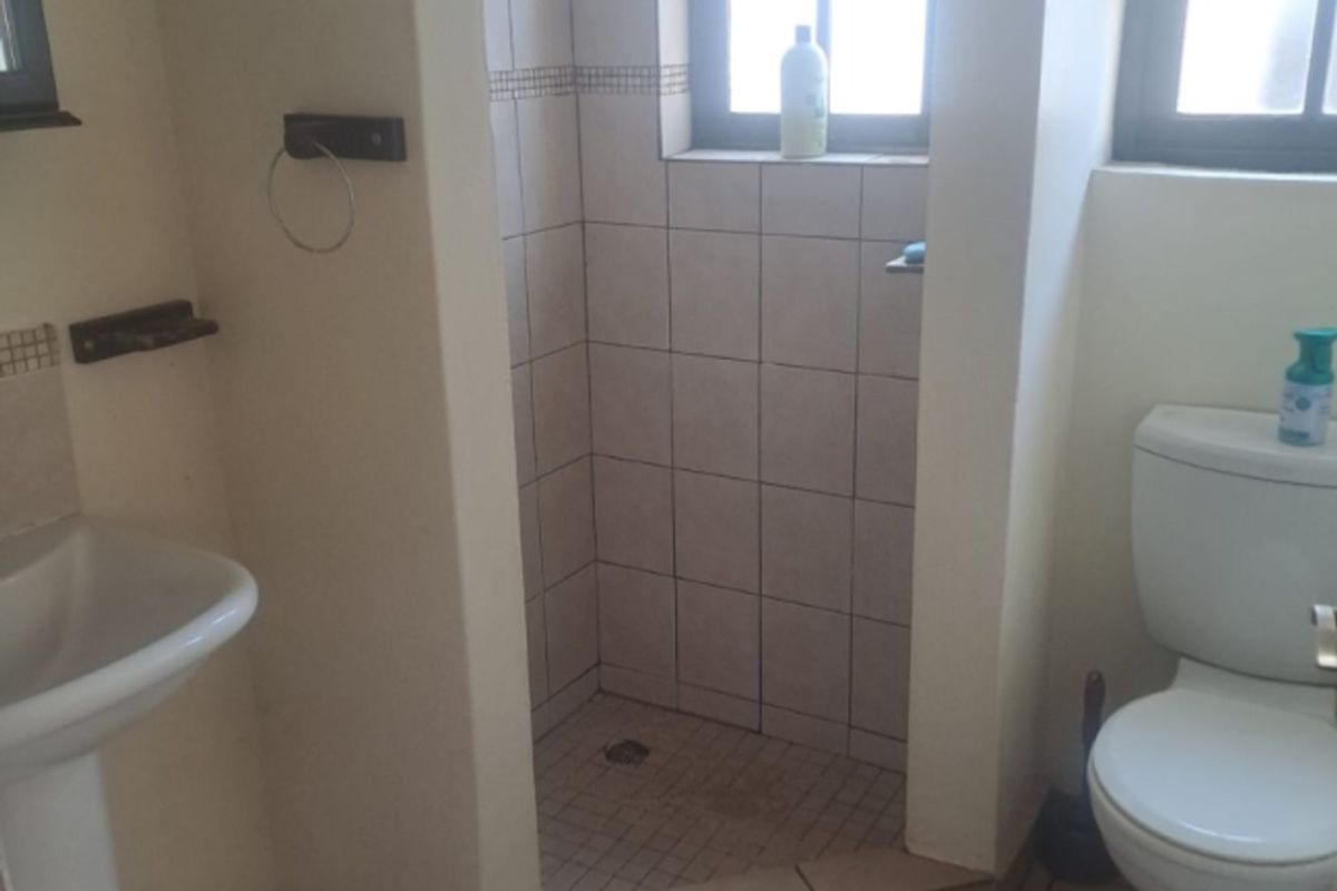 A functional bathroom featuring a pedestal basin, a tiled shower enclosure, and a standard toilet with tiled flooring and natural light from casement windows.