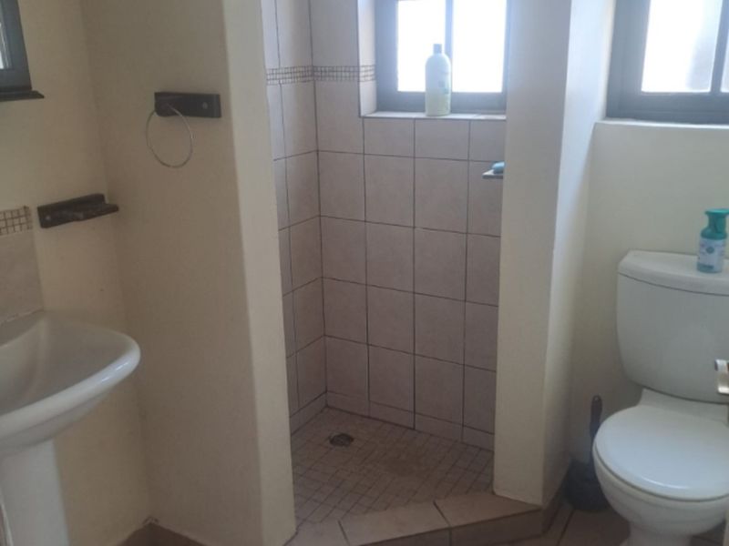 A functional bathroom featuring a pedestal basin, a tiled shower enclosure, and a standard toilet with tiled flooring and natural light from casement windows.