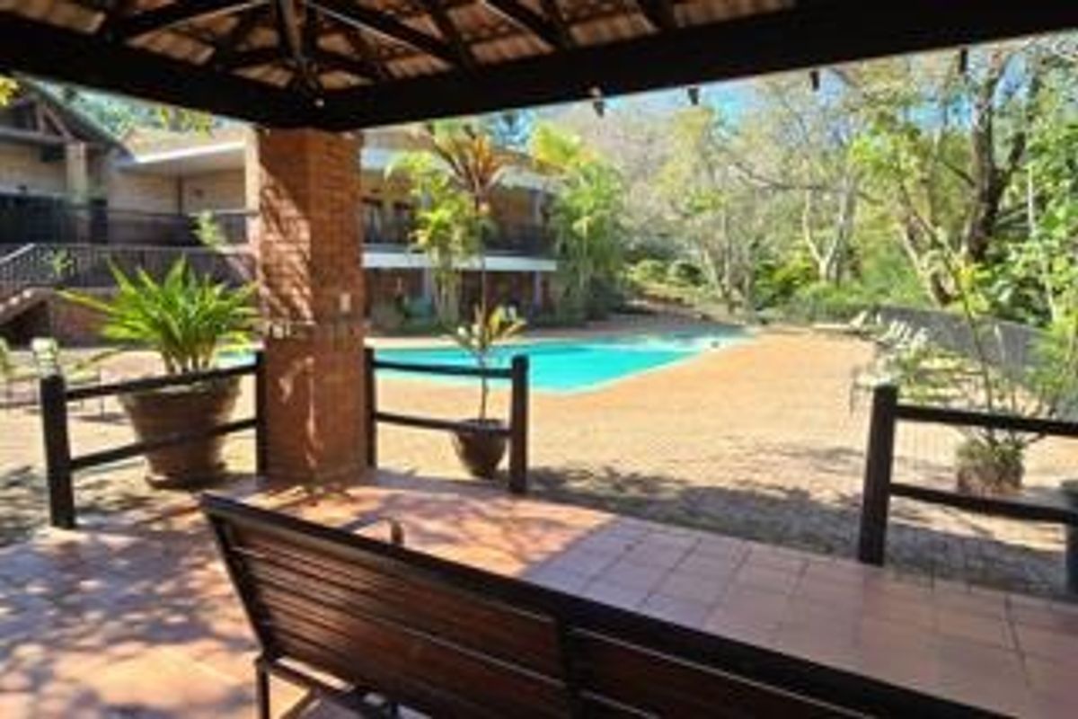A covered outdoor patio area overlooking a swimming pool and landscaped garden, featuring brick pillars and tiled flooring.