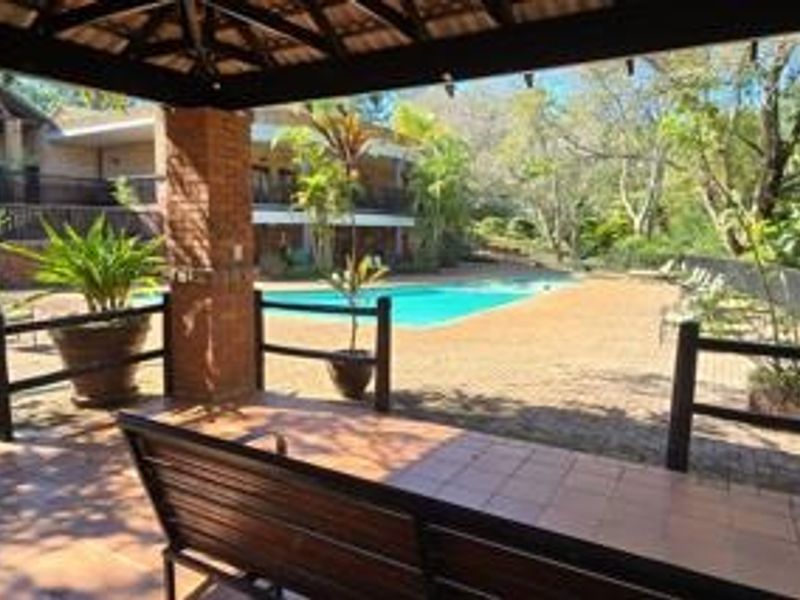 A covered outdoor patio area overlooking a swimming pool and landscaped garden, featuring brick pillars and tiled flooring.