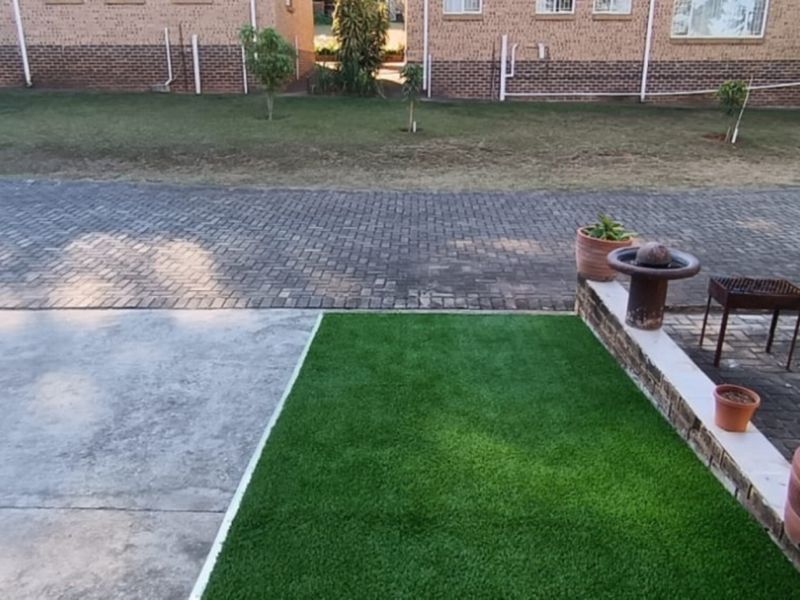 A view of the rear exterior area featuring a paved driveway, a section of artificial grass, and a low brick retaining wall.