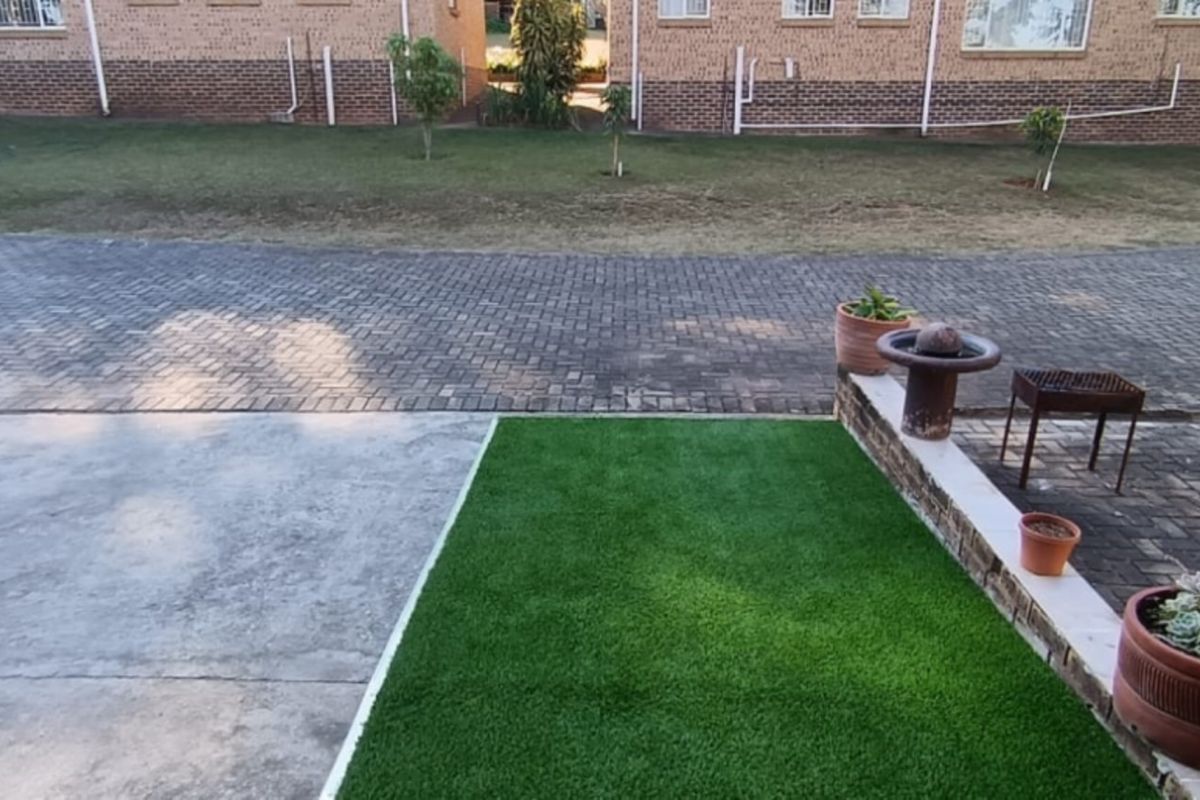 A view of the rear exterior area featuring a paved driveway, a section of artificial grass, and a low brick retaining wall.