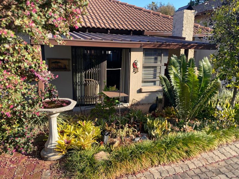 A well-maintained retirement village unit featuring a covered patio, brick chimney, and established garden with paved driveway access.