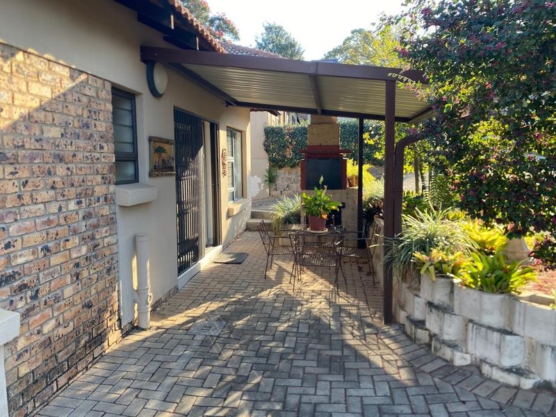 A paved outdoor patio area featuring a covered roof, built-in braai, and a retaining wall, providing a functional space for outdoor relaxation.