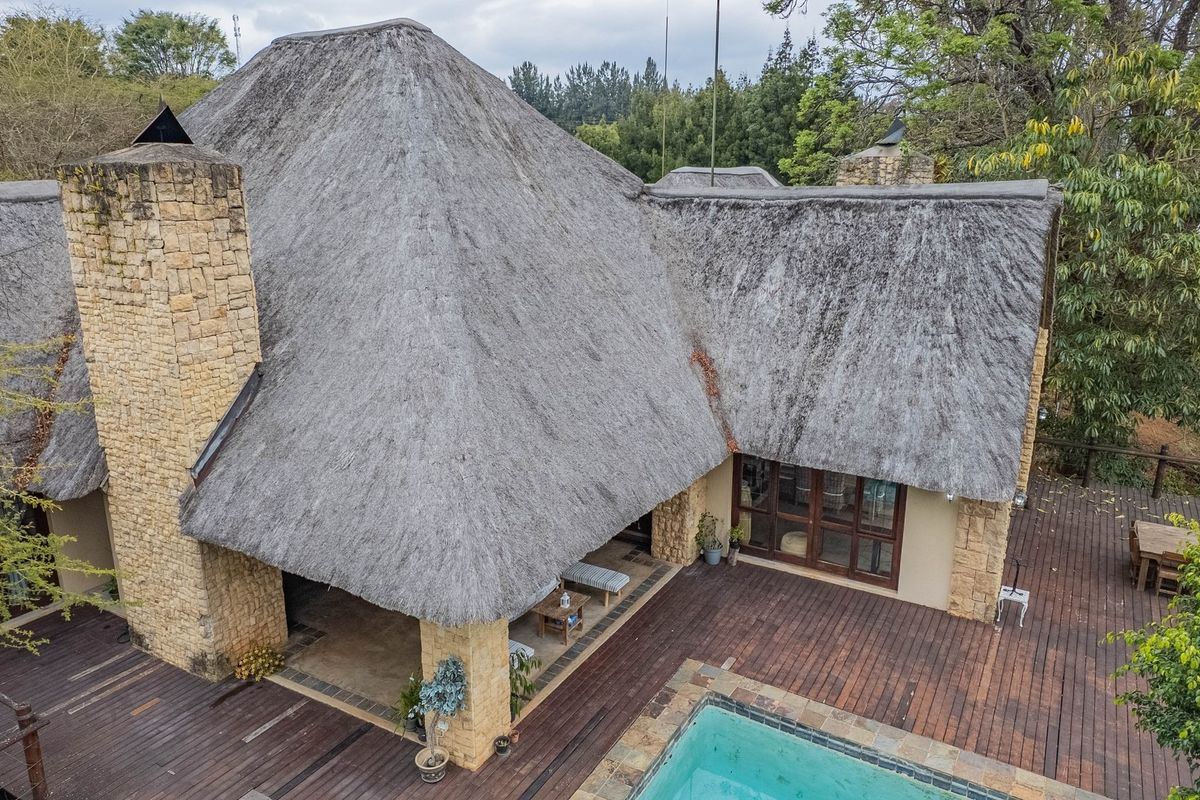Aerial view of a property featuring a large thatched roof, stone chimney, extensive timber decking, and a swimming pool set within a garden.