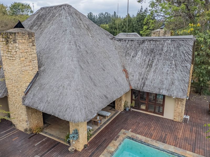 Aerial view of a property featuring a large thatched roof, stone chimney, extensive timber decking, and a swimming pool set within a garden.