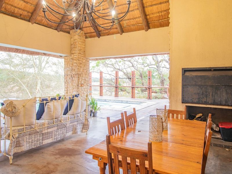 Spacious covered patio featuring a thatched roof, stone pillar, and built-in braai, overlooking the swimming pool and natural landscape.