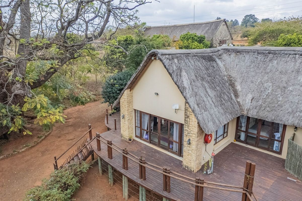 Aerial view of a thatched-roof cottage featuring a large timber deck, stone-clad exterior walls, and sliding glass doors set within a natural landscape.