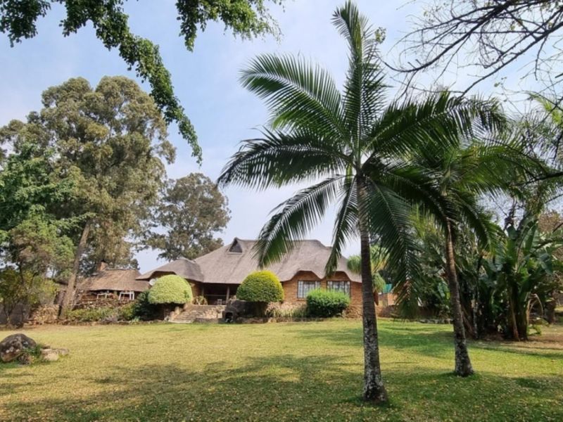 A view of the rear elevation of a thatch-roofed home set within a spacious, well-established garden with mature trees and stone accents.