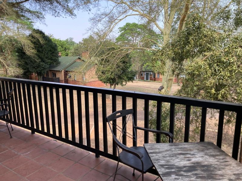A covered balcony featuring tiled flooring and a black railing, overlooking a garden area with mature trees and surrounding buildings.