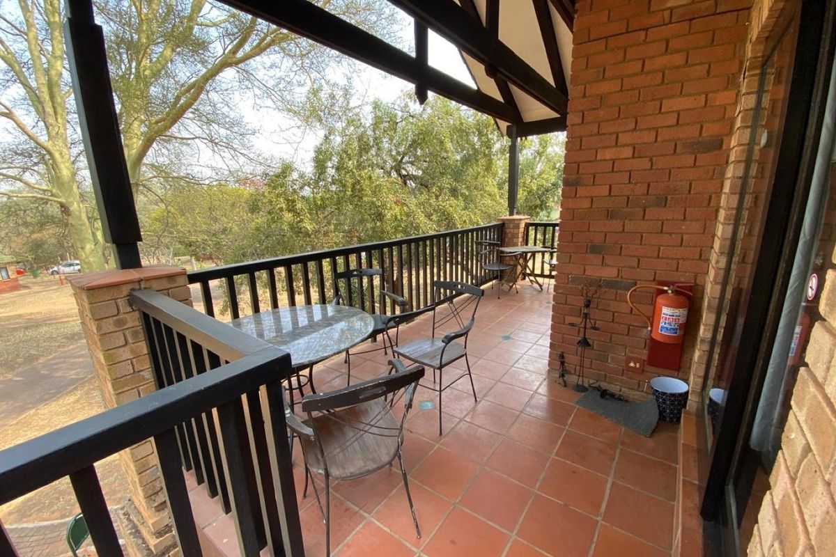 A covered balcony featuring exposed brick walls, tiled flooring, and a metal balustrade, offering an elevated outdoor space with views of the surrounding trees.