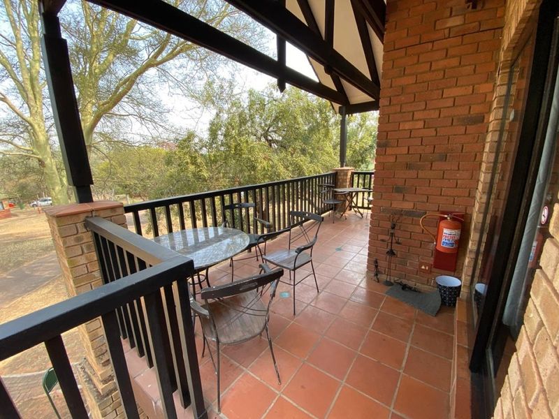 A covered balcony featuring exposed brick walls, tiled flooring, and a metal balustrade, offering an elevated outdoor space with views of the surrounding trees.