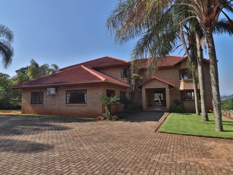 A well-maintained double-story brick house featuring a spacious paved driveway, covered entrance, and established garden surroundings.