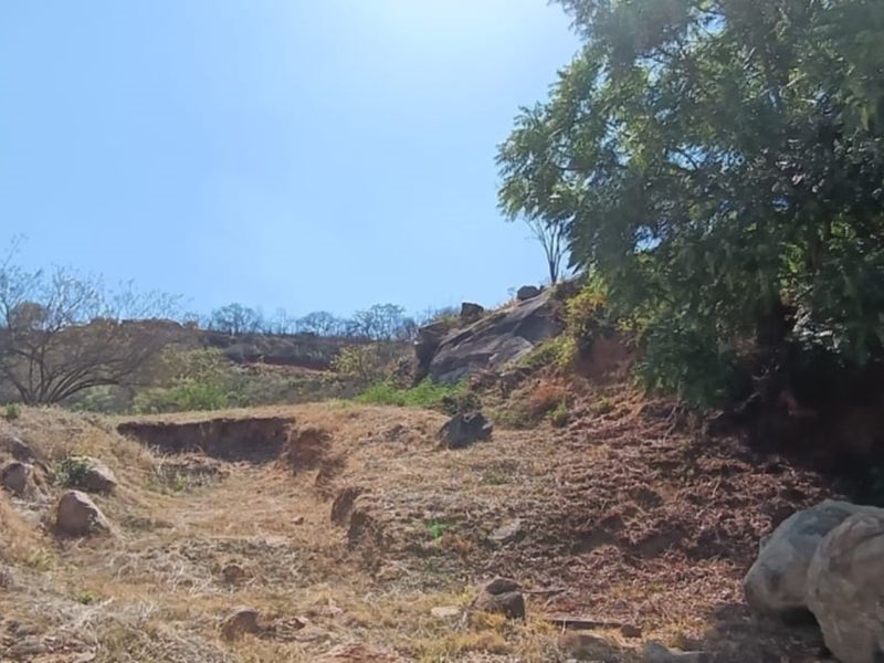A view of the vacant residential land featuring natural rock formations, mature trees, and sloping terrain under a clear sky.