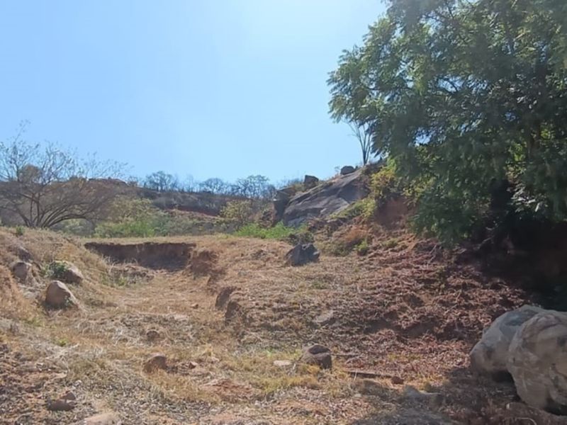 A view of a sloped, undeveloped residential plot featuring natural rock formations, mature trees, and visible soil erosion on the hillside.
