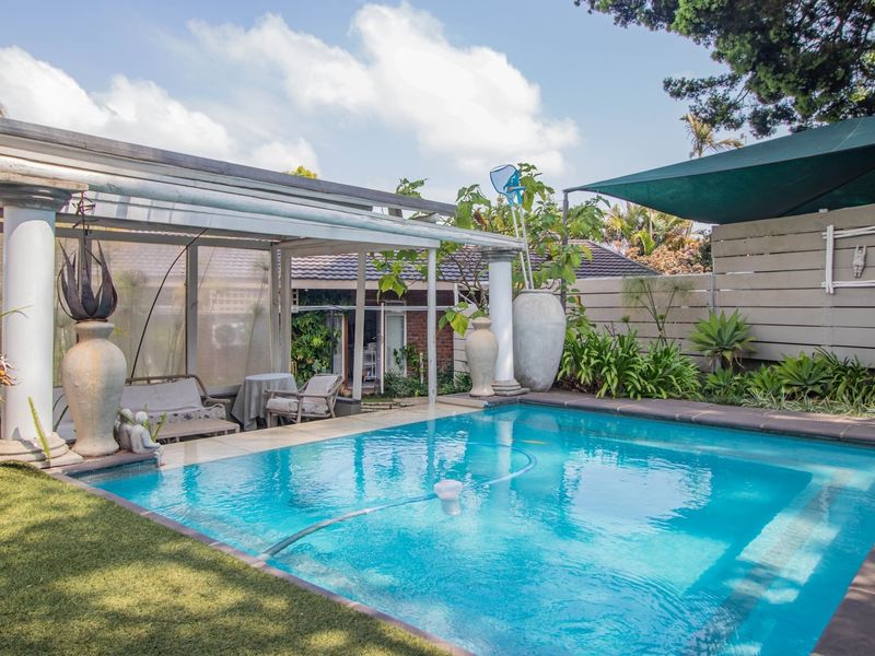 A well-maintained outdoor pool area featuring a covered patio with classical columns, a timber privacy screen, and a shade sail over the pool.