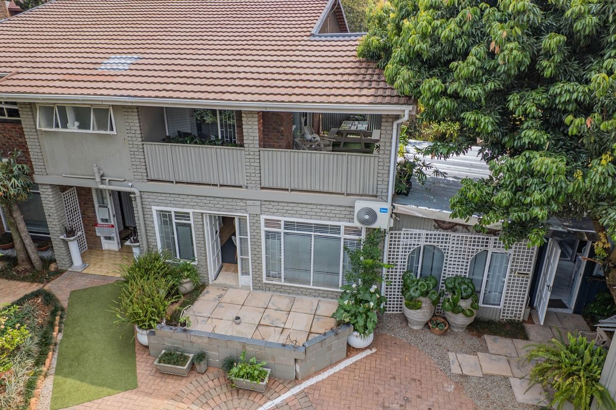Rear view of a two-story brick house featuring a balcony, ground-level patio, and paved garden area with mature trees.