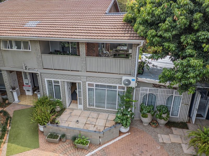 Rear view of a two-story brick house featuring a balcony, ground-level patio, and paved garden area with mature trees.