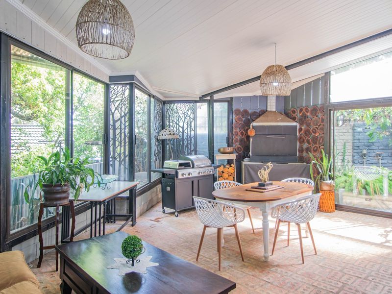 A bright, enclosed patio featuring a built-in braai, brick flooring, and expansive glass walls with decorative screens, offering a versatile indoor-outdoor entertainment space.