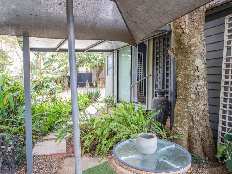 A covered patio area featuring a paved walkway, established garden surroundings, and direct access to the home through sliding glass doors.
