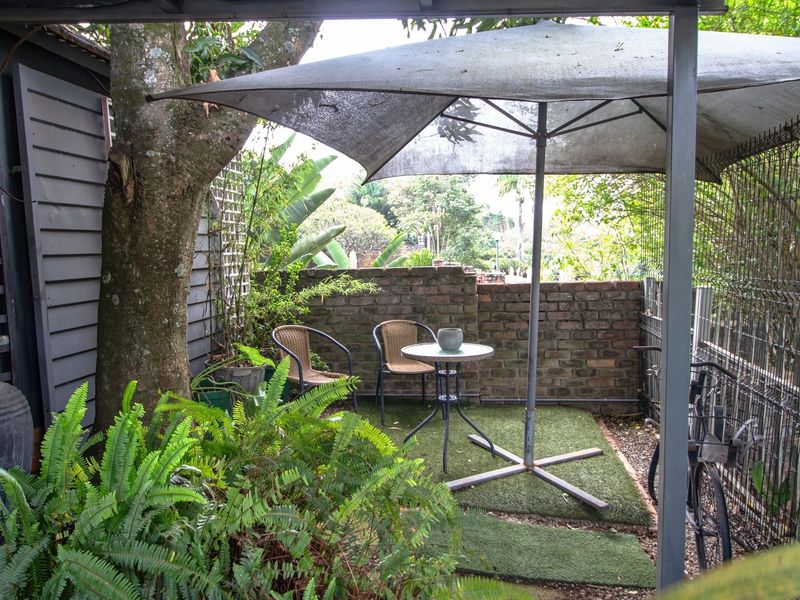 A compact outdoor patio area featuring a brick boundary wall, synthetic grass flooring, and a covered structure, providing a private space for relaxation.