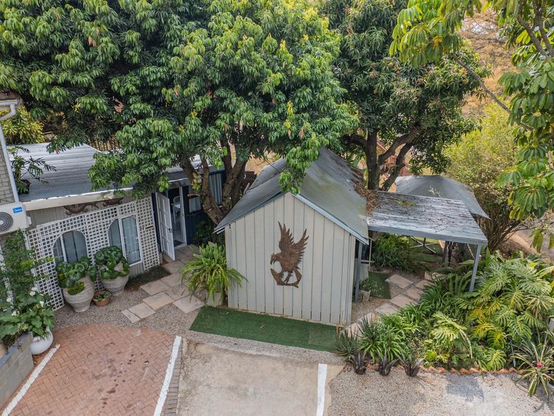 Aerial view of the rear exterior area featuring a paved driveway, metal-roofed structure, and mature trees providing shade over the garden space.