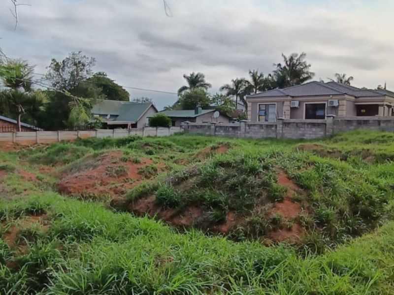 A vacant residential plot featuring uneven terrain and grass cover, enclosed by a perimeter wall in a suburban neighborhood setting.