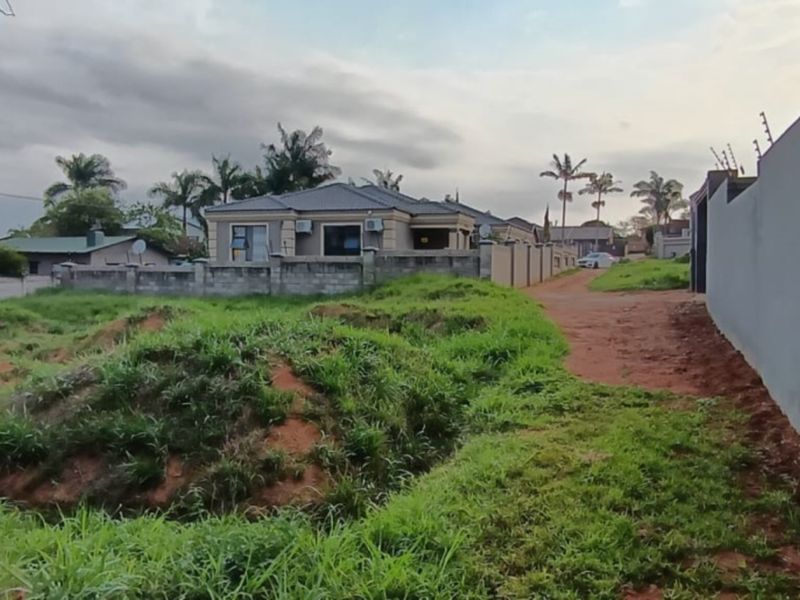 A vacant residential plot featuring an unpaved access path, perimeter walling with electric fencing, and uneven, grass-covered terrain.