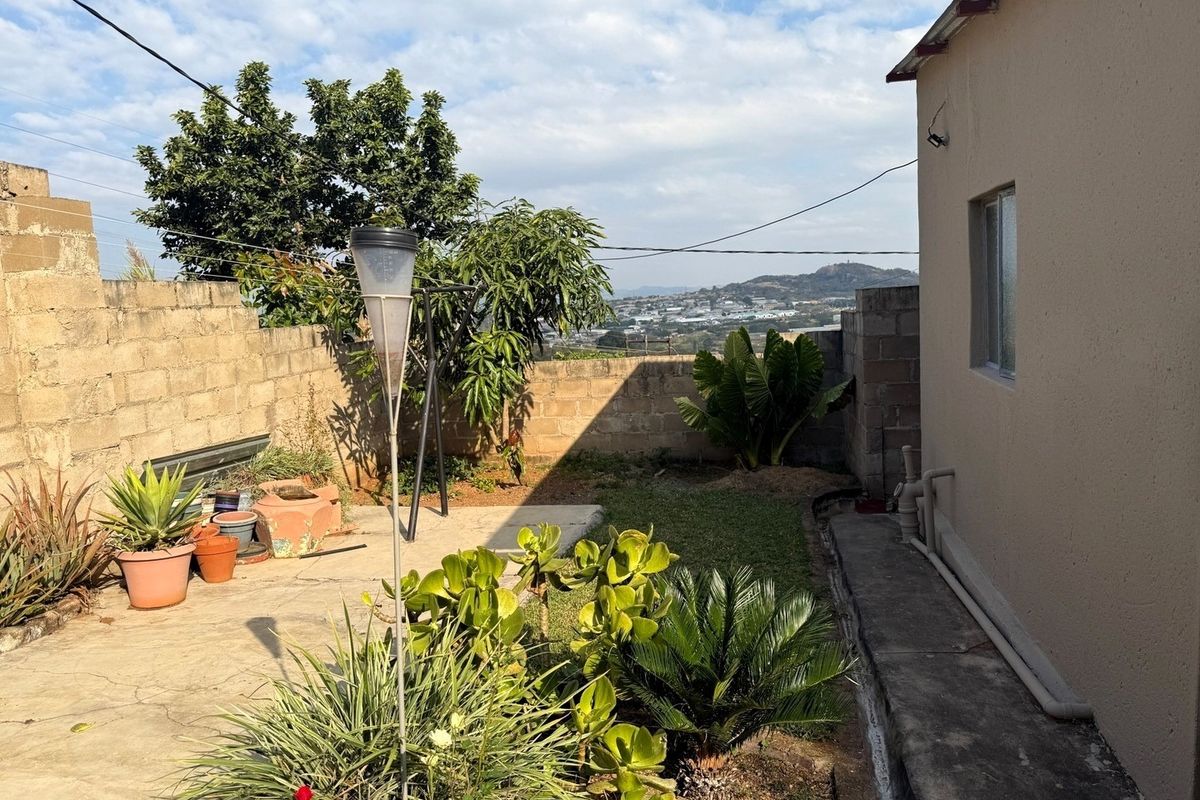 A rear exterior view of the property featuring a concrete paved area, perimeter block walling, and established garden landscaping with distant hillside views.