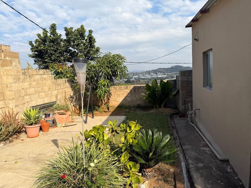 A rear exterior view of the property featuring a concrete paved area, perimeter block walling, and established garden landscaping with distant hillside views.