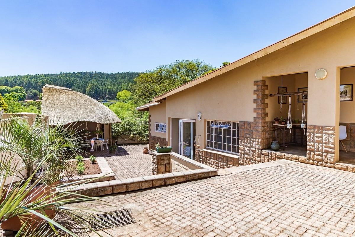 A spacious rear patio area featuring stone-clad walls, paved flooring, and a traditional thatched lapa, overlooking a scenic garden and forest landscape.