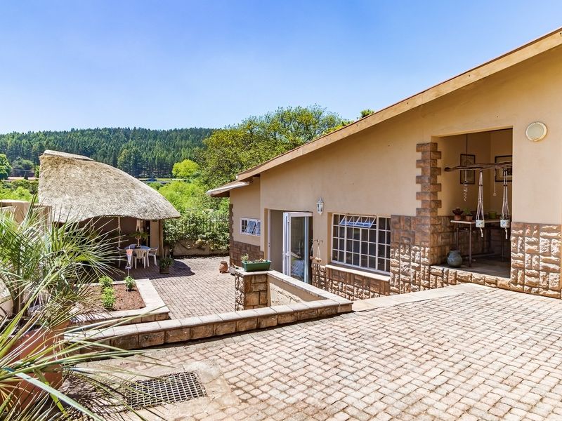 A spacious rear patio area featuring stone-clad walls, paved flooring, and a traditional thatched lapa, overlooking a scenic garden and forest landscape.