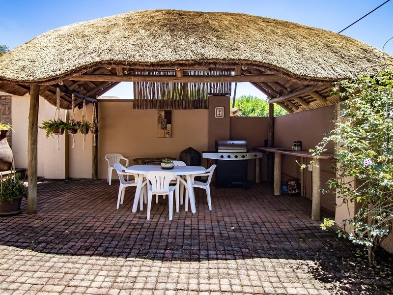 A covered outdoor patio area featuring a traditional thatched roof, timber support beams, brick paving, and a built-in braai station.