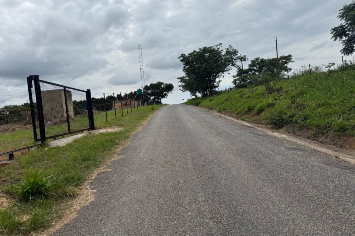 A view of the asphalt access road leading towards the property, featuring perimeter wire mesh fencing and utility infrastructure.