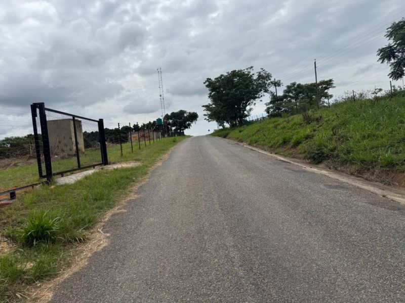A view of the asphalt access road leading towards the property, featuring perimeter wire mesh fencing and utility infrastructure.