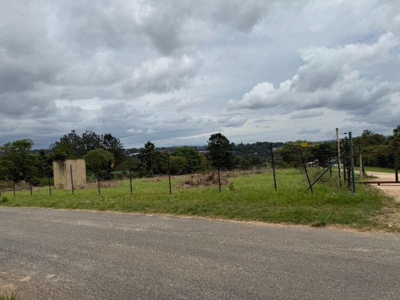 A vacant residential plot featuring an asphalt road frontage, electric fencing, and a small concrete structure, offering a clear view of the surrounding landscape.