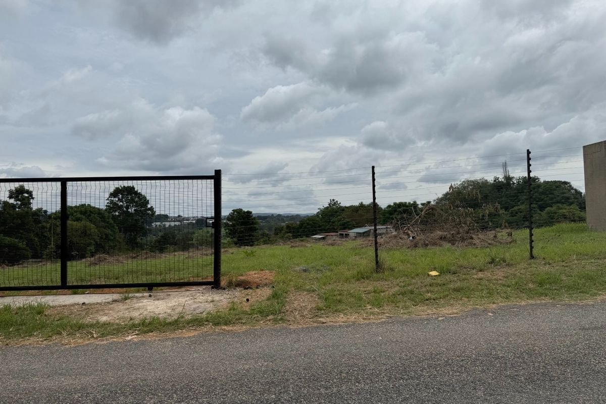 A vacant residential plot featuring a metal gate, electric fencing, and direct access from a paved road, offering an open landscape view.