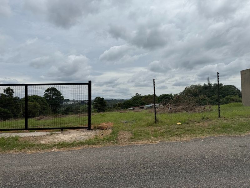 A vacant residential plot featuring a metal gate, electric fencing, and direct access from a paved road, offering an open landscape view.