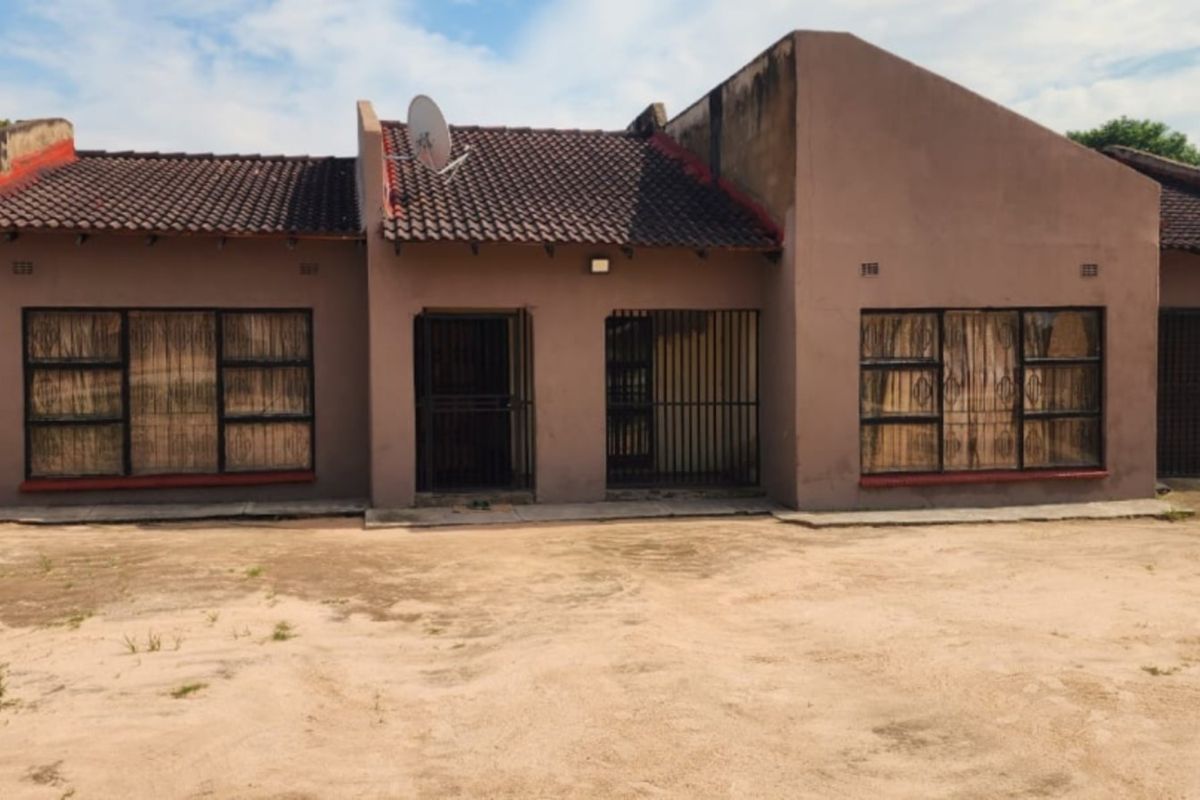 Front elevation of a single-story residential house featuring a tiled roof, security gates, and metal window frames, set on an unpaved plot.