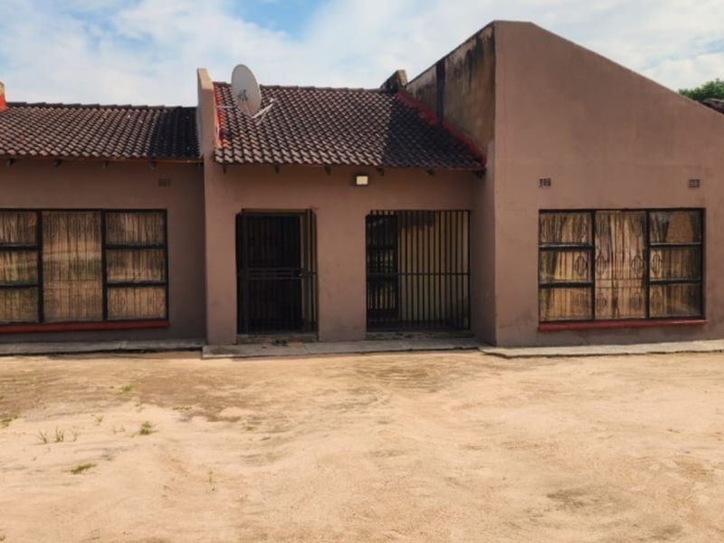 Front elevation of a single-story residential house featuring a tiled roof, security gates, and metal window frames, set on an unpaved plot.