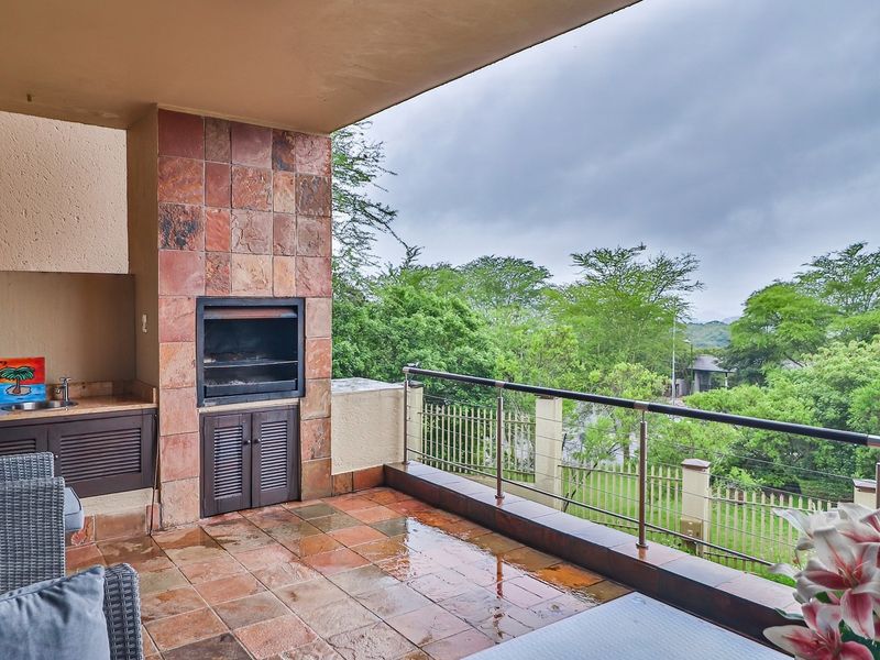 Covered balcony featuring a built-in braai, tiled flooring, and a stainless steel balustrade, offering pleasant views of the surrounding greenery.